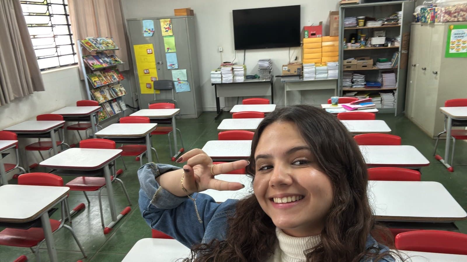 Isabela sorrindo dentro da sala de aula, com as carteiras vermelhas ao fundo